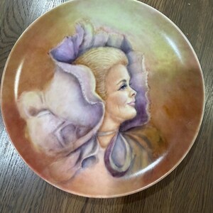 May include: A round decorative porcelain plate featuring a painted portrait of a person with blonde hair in profile. The individual is framed by a large, ruffled, light purple collar or garment. The background consists of blended warm brown and yellow tones.