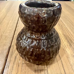 May include: A dark brown ceramic vase with a bulbous, hourglass-like shape and a textured, dimpled surface. The glaze has a glossy, mottled finish with subtle variations in tone, featuring a wide opening at the top and a rounded, wide base.