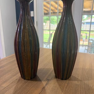 May include: Two tall, slender ceramic vases featuring a vertical ribbed texture. The vases have a multi-colored glaze with blended streaks of deep brown, forest green, and muted blue tones, finished with a narrow neck and a flared circular rim.