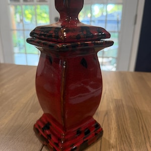 Puede incluir: Un recipiente de cerámica cuadrado con un esmalte rojo brillante y detalles negros moteados. El contenedor presenta una base acampanada, un cuerpo cónico y una tapa extraíble a juego con un asa cuadrada en la parte superior, todo con acabado liso.