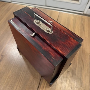 May include: A rectangular wooden box with a dark reddish-brown finish and black edges. The top features a silver-toned metal handle mounted on a rectangular plate, a small metal latch mechanism, and a thin silver-toned metal plaque on the side.