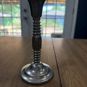 May include: A metallic candlestick holder with a dark, tarnished finish. The design features a wide, flared top cup, a slender central stem with multiple horizontal ribbed rings, a smooth rounded middle section, and a circular, flat base.
