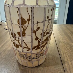 May include: A cylindrical vase featuring a mosaic design of cream-colored rectangular tiles with dark grout lines. The surface displays a brown painted branch motif with blossoms and black calligraphic text near the base, accented by red stamps.