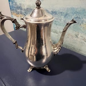 May include: A polished silver-toned coffee pot featuring an ornate handle, a curved spout with decorative detailing, and a lid topped with a finial. The vessel stands on three small, curved feet and has a smooth, reflective metallic surface.