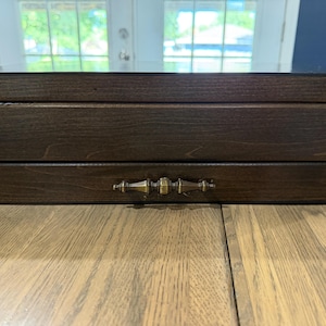 May include: A rectangular dark brown wooden storage box with a smooth finish. The box features a single horizontal drawer equipped with a decorative, elongated antique brass-toned metal handle centered on the front panel of the drawer.