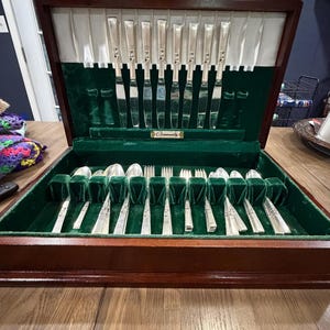 May include: A wooden flatware chest with a dark brown finish, featuring a green velvet interior lining. The set includes silver-toned knives stored in the lid and various spoons and forks arranged in the base. A small plaque reads Community.