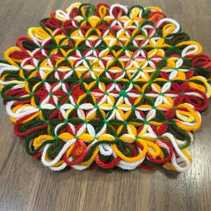 Könnte beinhalten: Ein runder, handgewebter Untersetzer aus Garn mit einem sich wiederholenden Blumenmuster in den Farben Dunkelgrün, leuchtendem Gelb, Weiß und Dunkelrot. Das Design besteht aus ineinandergreifenden Garnschlaufen mit strukturierter Oberfläche.