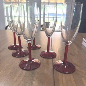 May include: A set of six clear glass champagne flutes, each featuring a slender stem and a circular base in a deep red color. The glasses have a tall, narrow bowl design, providing a consistent look across the entire collection of drinkware.