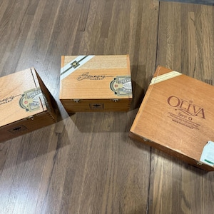 May include: Three rectangular wooden cigar boxes with hinged lids and metal latches. Two boxes feature Genesis Habano branding with white and green diagonal labels. The third box is labeled Oliva Serie O with black text and a white corner seal.