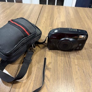 May include: A black Canon Sure Shot 20m point-and-shoot film camera with a lens, flash, and digital display, positioned next to a black rectangular camera bag featuring a red horizontal stripe and an adjustable black shoulder strap.
