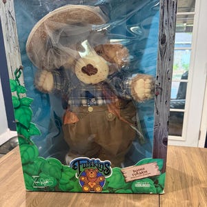 May include: A Boone Furskin plush bear doll by Coleco in its original packaging. The bear wears a blue plaid shirt, brown pants with suspenders, and a beekeeper hat with netting. The box features green leaf graphics and the text Furskins.