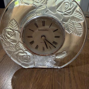 May include: A clear glass desk clock featuring a circular face with black Roman numerals and the text HOME BEAUTIFUL. The glass frame is decorated with frosted, raised rose and leaf motifs. The clock has three black hands and a silver-toned bezel.