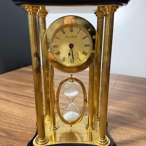 May include: A gold-tone metal and black wood desk clock featuring a circular clock face with Roman numerals and the text Bey-Berk. Below the clock is a glass hourglass with white sand. A plaque reads Friends are timeless, Intrust Bank N.A., August 31 2005.