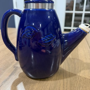 May include: A glossy cobalt blue ceramic oil cruet featuring a curved handle, a tapered spout with a white plastic stopper, and a metallic silver band around the neck. The body of the cruet is decorated with embossed leaf patterns on the front.