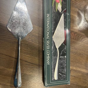 May include: A silver-plated cake server with an ornate floral pattern on the triangular blade and a smooth handle, accompanied by its original dark green box labeled International Silver Company, which states it is 11.25 inches in length.