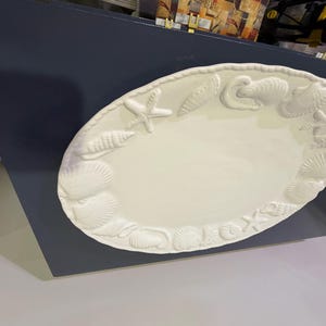 May include: An oval-shaped cream-colored ceramic platter featuring a raised, embossed border decorated with various seashells and starfish. The center of the platter is smooth and plain, providing a contrast to the textured, detailed rim.