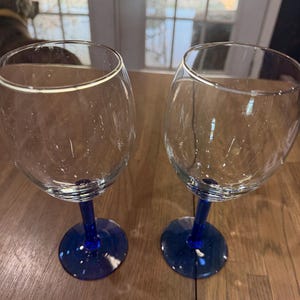 May include: Two clear glass wine glasses featuring deep blue stems and circular blue bases. The bowls of the glasses are transparent and rounded, transitioning into slender blue stems that connect to solid, flat blue circular bases.
