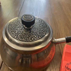 May include: A stainless steel pot with a rounded body and a flat lid featuring an intricate embossed floral pattern. The lid has a central black knob with a small floral detail, and the pot includes a side handle with a black grip.