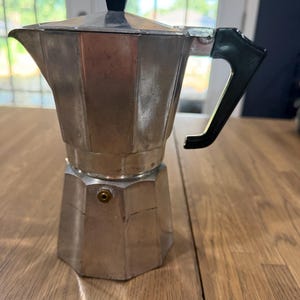 May include: A silver-colored aluminum stovetop espresso maker with an octagonal base and body. It features a black plastic handle and a matching black knob on the hinged lid. A small brass-colored pressure release valve is visible on the lower chamber.
