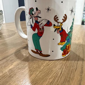 May include: A white ceramic mug featuring a colorful illustration of Goofy wearing a red sweater and green pants, alongside Pluto wearing reindeer antlers and a red sweater. The design includes a Christmas tree with gifts and black star accents.