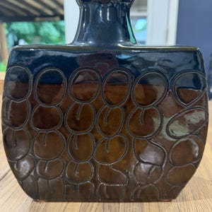 May include: A flat, rectangular ceramic vase with a glossy finish. The top section is a deep, solid black, while the main body features a brown glaze with a repeating, embossed pattern of rounded, pebble-like shapes outlined in dark, recessed lines.
