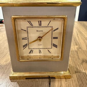 May include: A rectangular Bulova quartz clock featuring a silver-toned body with gold-toned trim at the top and bottom. The square clock face has a cream background, black Roman numerals, and gold-toned hands. The word Bulova is printed in black.