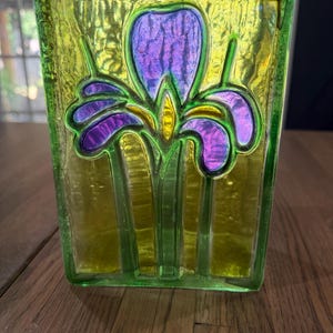 May include: A rectangular glass vase featuring a textured surface with a vibrant yellow background and a central iris flower motif in shades of purple and yellow. The glass has a translucent quality with raised outlines defining the floral design.
