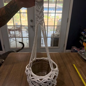 Peut inclure: Une suspension pour plantes en macramé blanc dotée d'un anneau en bois circulaire au sommet et d'une structure en corde tressée qui soutient un récipient rond. La suspension est maintenue par plusieurs cordes nouées formant un motif en treillis.