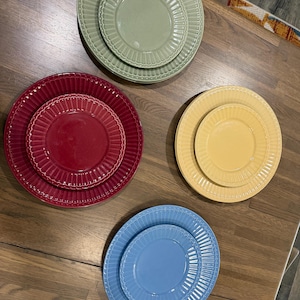 May include: Four sets of round ceramic plates in sage green, deep red, sunny yellow, and sky blue. Each set includes a larger dinner plate and a smaller salad plate, both featuring a raised, fluted, and beaded rim design with a smooth center.