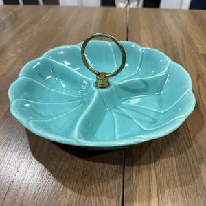 May include: A turquoise ceramic serving dish with a scalloped, flower-like shape divided into three distinct sections. A central brass-toned metal ring handle is attached to the middle, providing a convenient way to lift the dish.