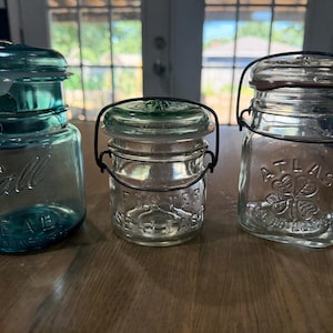 Pode incluir: Três frascos de vidro vintage com fechos de arame. O frasco da esquerda é de vidro azul com o texto em relevo Ball IDEAL. O frasco central é de vidro transparente com o texto FOSTER SEALFAST. O frasco da direita é de vidro transparente com o texto ATLAS e um ícone de trevo.