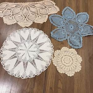 May include: Four handmade crochet doilies in various shapes and sizes. One is a large beige semi-circle, one is a light blue six-pointed star shape, one is a large circular white piece with a starburst pattern, and one is a small round beige doily.