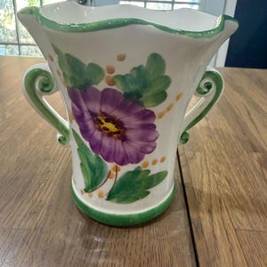 May include: A white ceramic vase with a scalloped rim and two side handles. The exterior features a hand-painted purple flower with a yellow center, surrounded by green leaves and small yellow dots. A green band accents the base and rim.