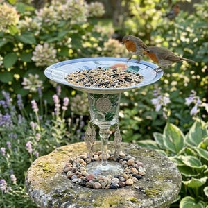 May include: A bird feeder constructed from a shallow ceramic plate with a floral pattern, supported by a clear glass pedestal filled with green glass marbles and adorned with hanging silver leaf charms, filled with a mix of bird seeds.