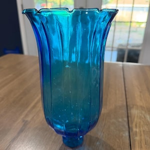 May include: A vibrant blue glass hurricane candle holder featuring a vertical ribbed texture and a flared, scalloped rim. The glass exhibits a translucent quality with a slight gradient, transitioning from a lighter cyan hue at the top to a deeper blue base.