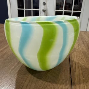 May include: A rounded glass bowl featuring a repeating pattern of vertical, wavy stripes in alternating shades of lime green, light blue, and opaque white. The bowl has a smooth, polished finish and a wide, open top with a slightly irregular rim.