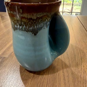 May include: A ceramic mug featuring a light blue base color that transitions into a dark brown glaze at the rim. The mug has a unique, integrated hand-warming pocket handle design that is molded directly into the side of the vessel body.