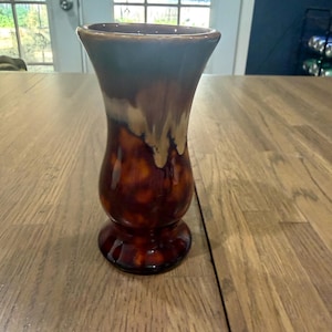 May include: A ceramic vase featuring a flared rim and a bulbous base. The glaze transitions from a dark, glossy brown at the bottom to a lighter, creamy tan in the middle, and finishes with a deep, muted blue-grey tone at the top opening.