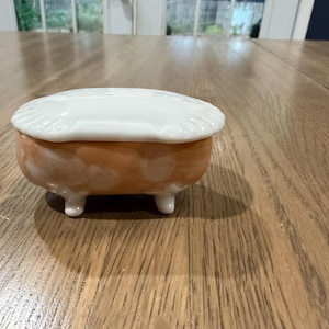 Pode incluir: Uma pequena caixa de cerâmica de formato oval, concebida para se assemelhar a uma banheira em miniatura com pés. A base tem uma cor pêssego e branca manchada, enquanto a tampa removível é branca sólida com uma borda recortada.