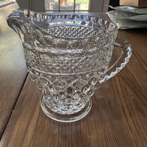 May include: A clear glass pitcher featuring a prominent diamond-cut pattern across its body. The vessel includes a wide pouring spout, a sturdy handle with a scalloped edge, and a circular base. The glass is transparent with a reflective surface.