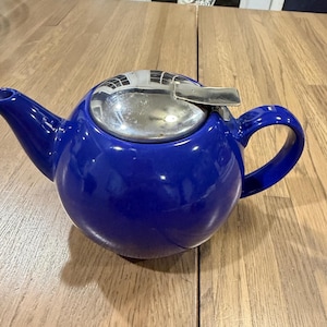 May include: A round, cobalt blue ceramic teapot featuring a polished, reflective silver-tone metal lid with a hinged handle. The teapot has a curved spout and a sturdy side handle, showcasing a smooth, glossy finish throughout the main body.