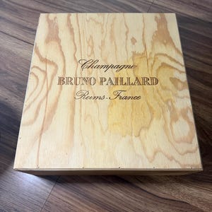 May include: A rectangular light-colored wooden box with a natural grain pattern. The lid features the dark engraved text "Champagne", "BRUNO PAILLARD", and "Reims-France" centered on the surface. The box has a smooth finish and clean edges.