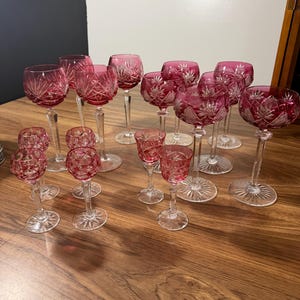 May include: A collection of fifteen cranberry red and clear cut crystal glasses of varying sizes and shapes. Each glass features intricate starburst and geometric patterns etched into the colored bowls, supported by clear stems and circular bases.