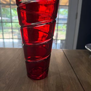 Peut inclure: Un vase en verre rouge vif présentant un design en spirale distinct qui crée une série de segments arrondis empilés. Le verre est translucide, laissant passer la lumière à travers la couleur cramoisie profonde, avec un sommet large et ouvert.