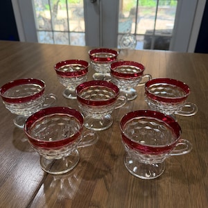 May include: A set of eight clear glass cups with a textured diamond pattern and a solid red rim. Each cup features a short pedestal base and a small handle, arranged in a group on a wooden surface. The glass has a faceted design throughout.