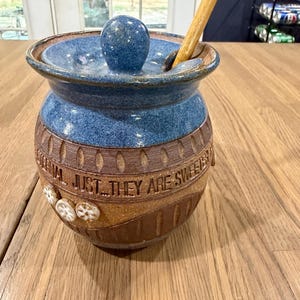 May include: A ceramic honey pot with a blue glazed lid and a matching blue upper section. The lower body is brown with a textured band featuring the embossed text "JUST... THEY ARE SWEETER" and small white flower accents. A wooden honey dipper is included.
