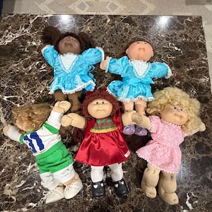 May include: Five dolls with yarn hair and soft bodies. Two wear bright blue dresses with white lace trim. One wears a green and white athletic outfit. One wears a red dress with a green plaid bodice. One wears a pink floral dress with white lace trim.