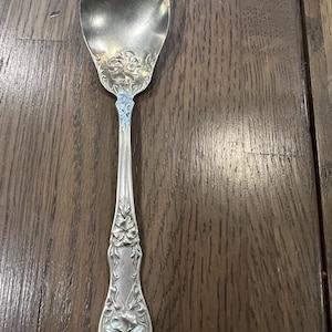 May include: A silver-toned metal spoon featuring an ornate floral relief pattern on the handle and the base of the bowl. The design includes detailed embossed flower motifs and leaf accents that extend from the bottom of the handle upward.