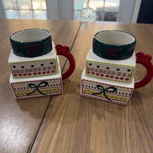 May include: Two ceramic mugs shaped like stacked gift boxes. Each mug features a dark green rim, a red handle, and a white body decorated with red and green patterns of dots, dashes, and bows. The mugs have a square base and a cylindrical top.