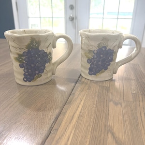 May include: Two off-white ceramic mugs with a textured, wavy surface. Each mug features a painted cluster of dark purple grapes with green leaves and thin, brown vine tendrils. The mugs have a sturdy handle on the right side and a flared rim.